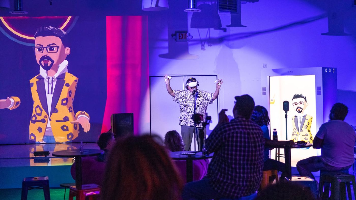 A comic in a VR headset on the live stage while his avatar is projected on the wall