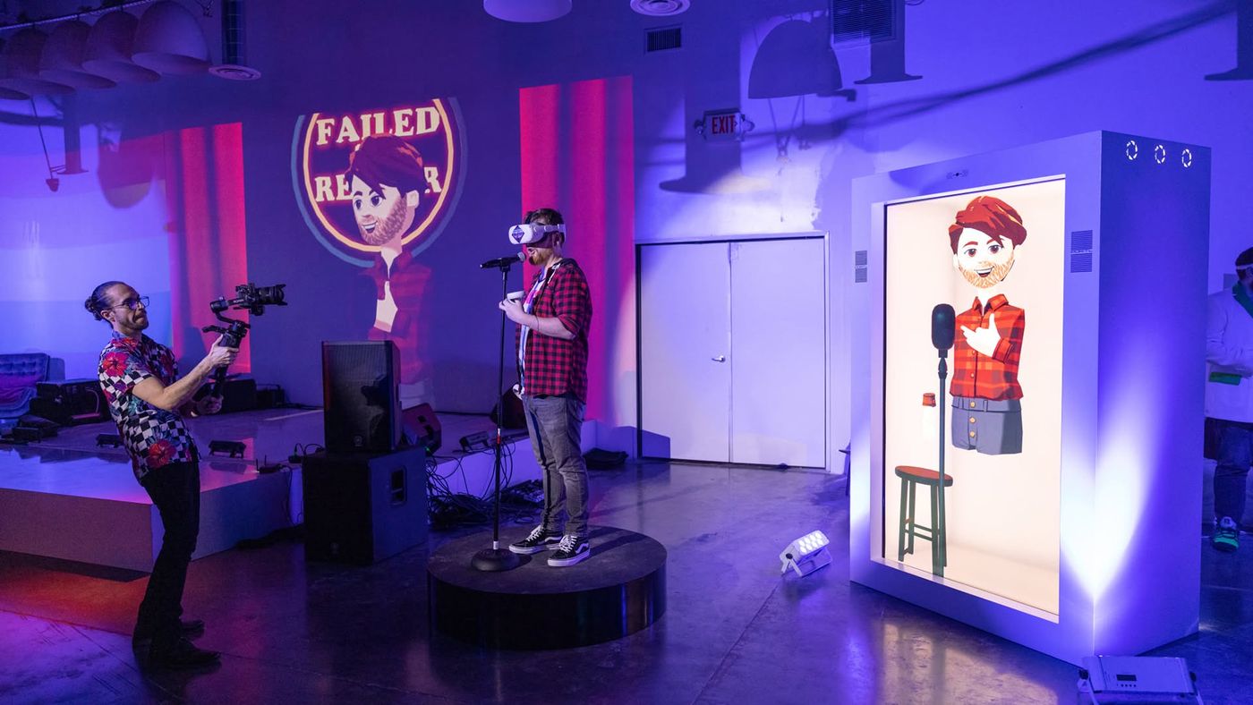 A comic in a VR headset on stage, his avatar in the Proto hologram box beside him